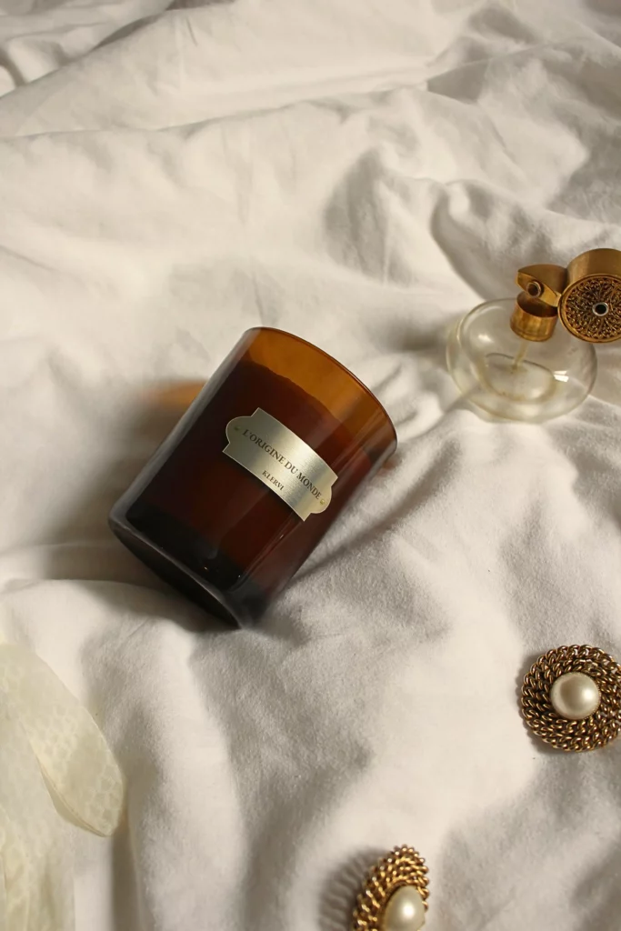 imagine d'une bougie parfumée de la marque Klervi Paris, posée sur un joli drap blanc et entourée d'un flacon de parfum et de belles boucles d'oreilles